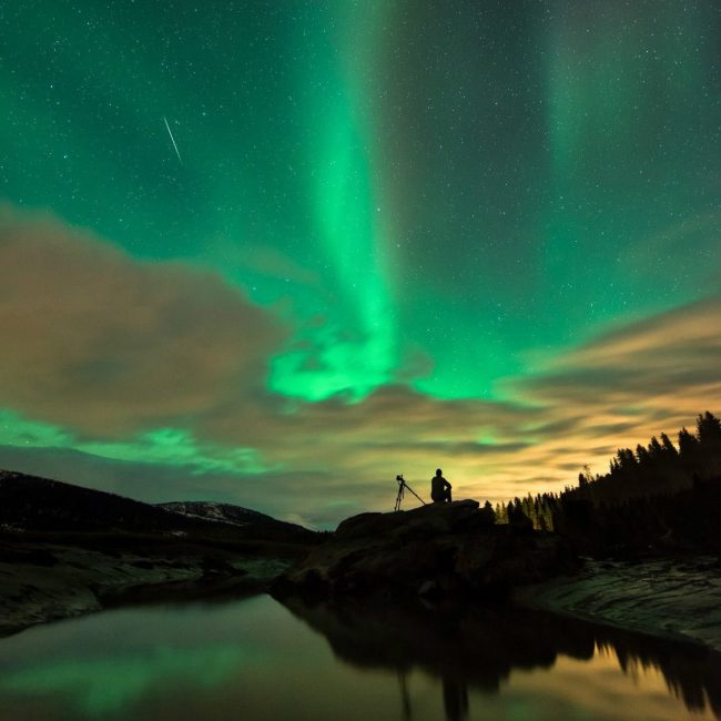 A man with a camera tripod shadowed watching a thin line in the sky against a giant green aurora.